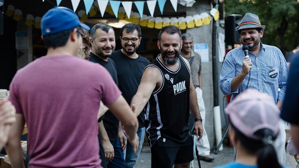 Eine Gruppe von Menschen lächelt und schüttelt sich die Hände bei einem Treffen im Freien mit blau-gelben Wimpeln und frischen Brezeln.