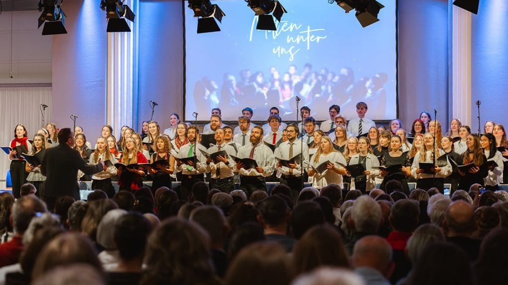 Ein Chor tritt auf der Bühne vor einem Publikum auf, mit einem Dirigenten an der Spitze und einer Leinwand, die Heaven Winter Uns zeigt.