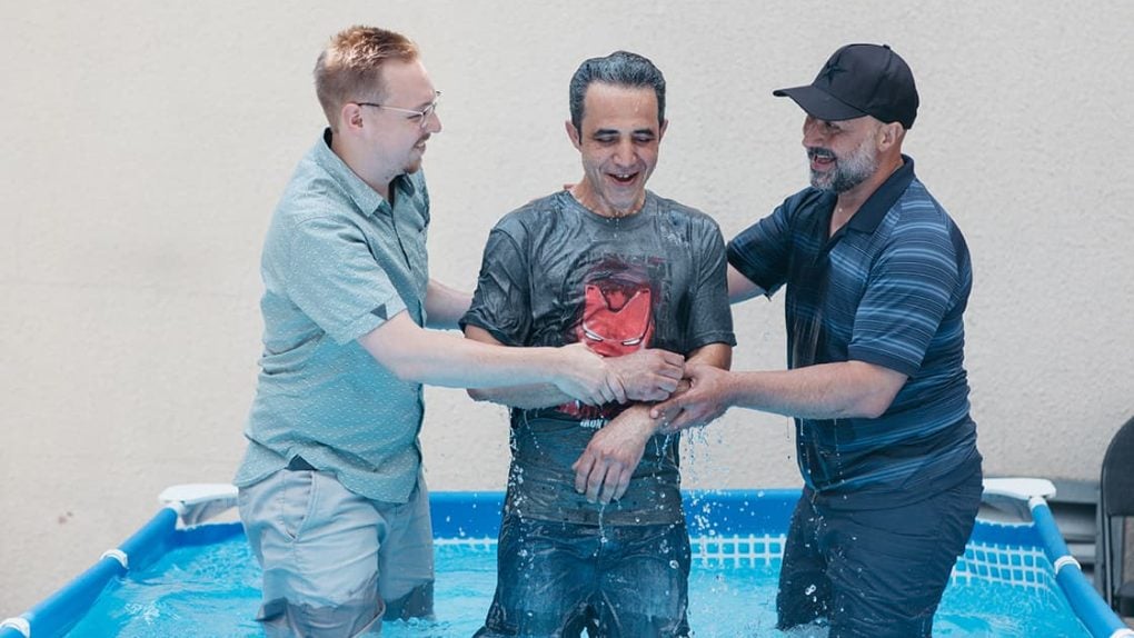 Drei Männer stehen lächelnd in einem kleinen Becken, wobei zwei Männer dem dritten Mann in der Mitte helfen.