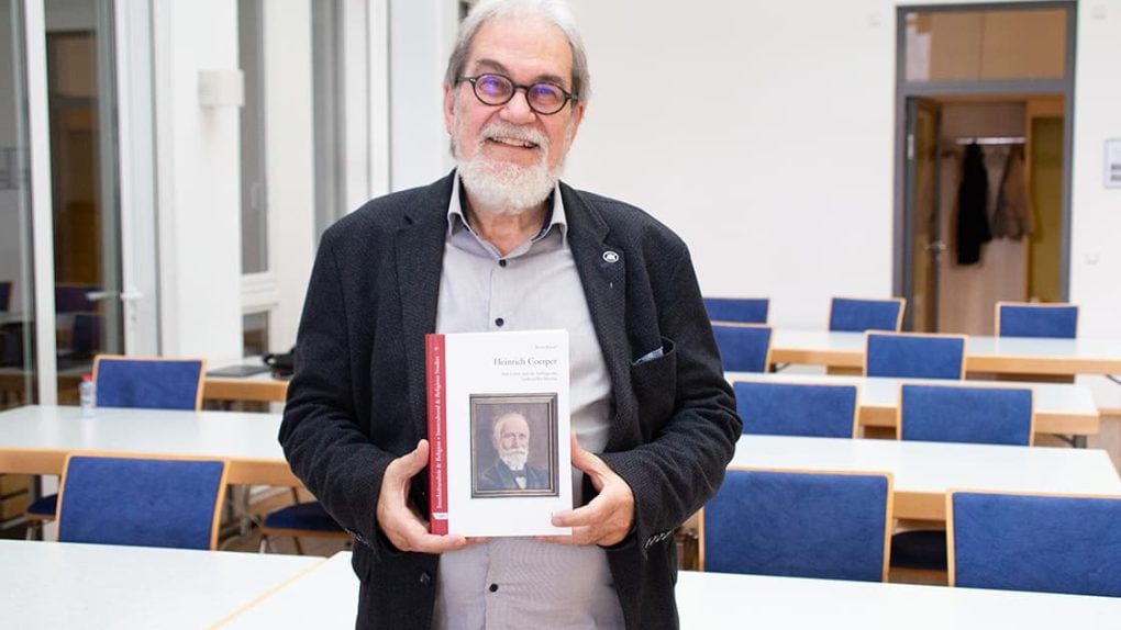 Ein lächelnder Mann mit Brille hält ein Buch mit einem Porträt auf dem Einband in einem Klassenzimmer mit blauen Stühlen und weißen Wänden.