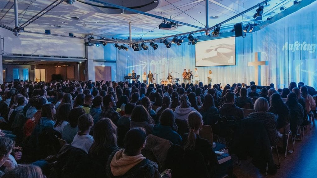 Ein großes Publikum schaut einer Live-Band zu, die in einer hell erleuchteten Halle mit blauer Beleuchtung auftritt.