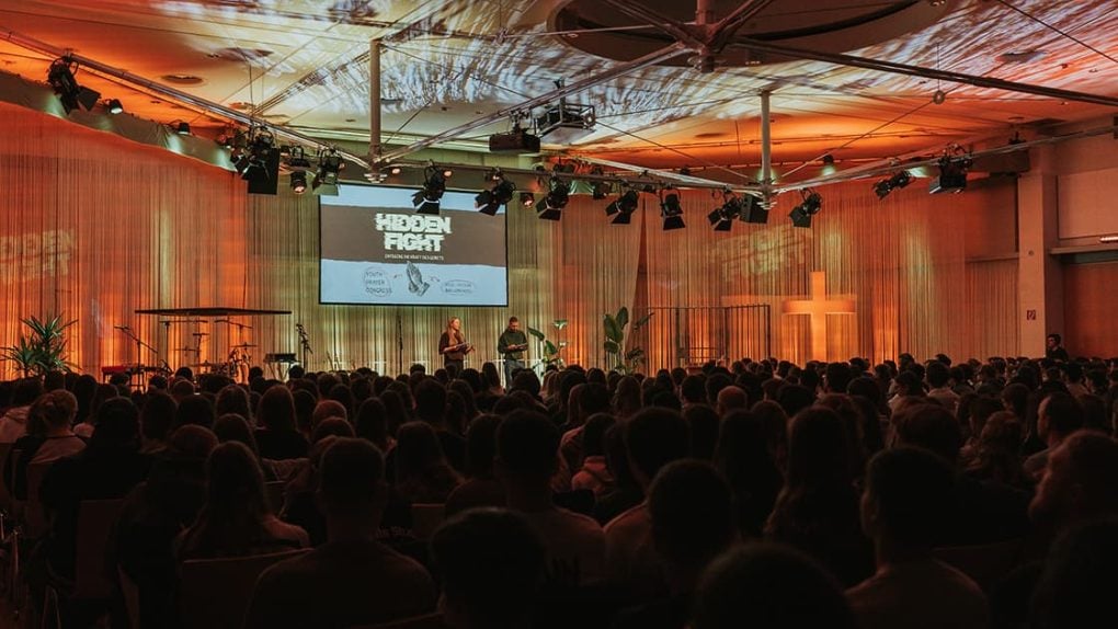 Ein großes Publikum verfolgt eine Bühnenpräsentation mit dem Titel Hidden Fight in einem warm beleuchteten Auditorium mit einem Kreuz an der Wand.