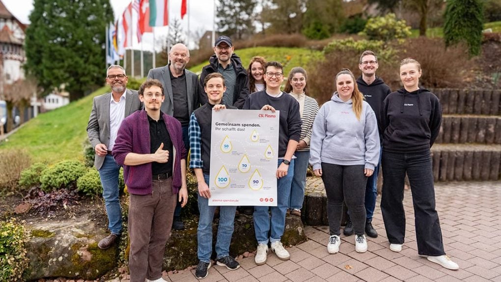 Eine Gruppe von elf Personen steht im Freien, lächelt und hält ein Plakat mit Texten und Diagrammen über HIV und Blutspenden.