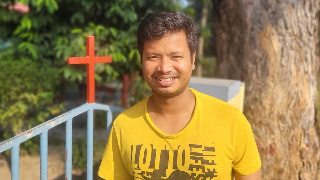 Ein lächelnder Mann in einem gelben Hemd steht vor einem Tor mit einem roten Kreuz und einem großen Baum im Hintergrund.