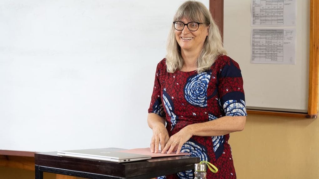 Eine lächelnde Frau mit langen grauen Haaren steht an einem Schreibtisch vor einem Whiteboard, trägt eine Brille und ein rot gemustertes Kleid.