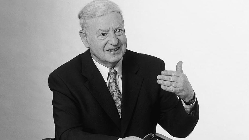 Älterer Mann im Anzug sitzt und gestikuliert mit seiner rechten Hand, in der linken Hand hält er eine Brille, vor einem schlichten Hintergrund.