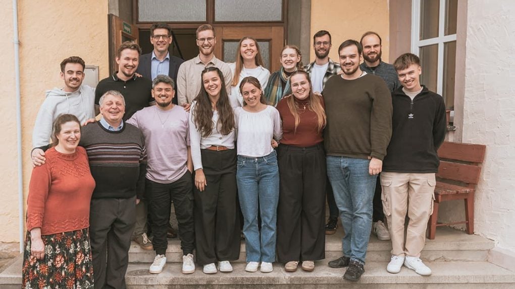 Fünfzehn lächelnde Menschen stehen auf einer Freitreppe vor einem Gebäude für ein Gruppenfoto zusammen.