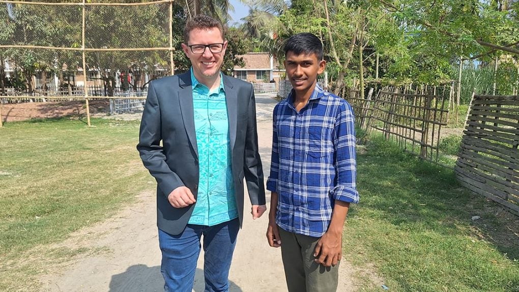 Zwei Männer, einer im Blazer, der andere im karierten Hemd, stehen auf einem sonnigen Weg, der von Grünflächen und Zäunen umgeben ist.