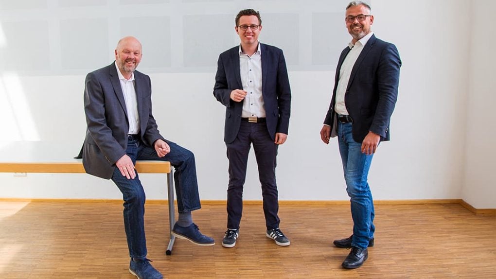 Drei Männer in legerer Geschäftskleidung posieren und lächeln in einem hellen, minimalistischen Raum mit Holzboden und weißen Wänden.