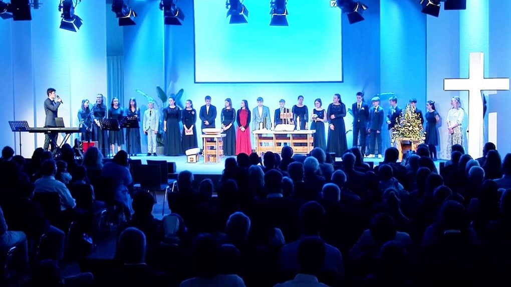 Ein Chor steht in einer Kirche auf der Bühne, dem Publikum zugewandt, mit blauer Beleuchtung und einem großen beleuchteten Kreuz.