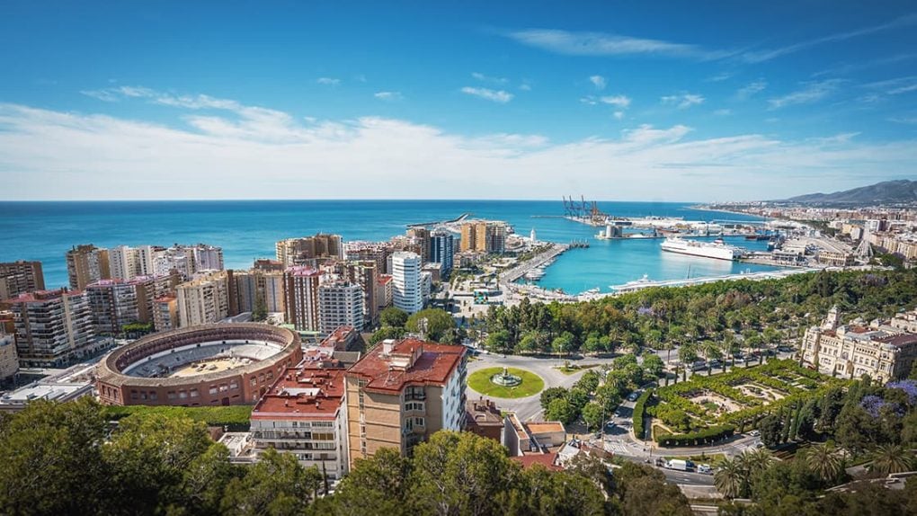 Luftaufnahme einer Küstenstadt mit einer Stierkampfarena, Hochhäusern, einem Jachthafen und dem Meer unter blauem Himmel.
