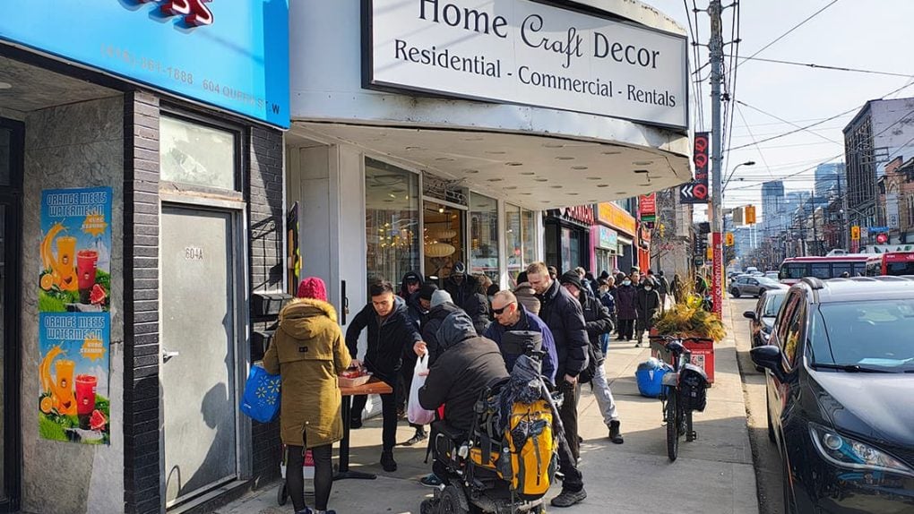 An einem sonnigen Tag versammeln sich Menschen um einen Tisch auf einem belebten Bürgersteig vor einem Geschäft für Heimwerkerbedarf.