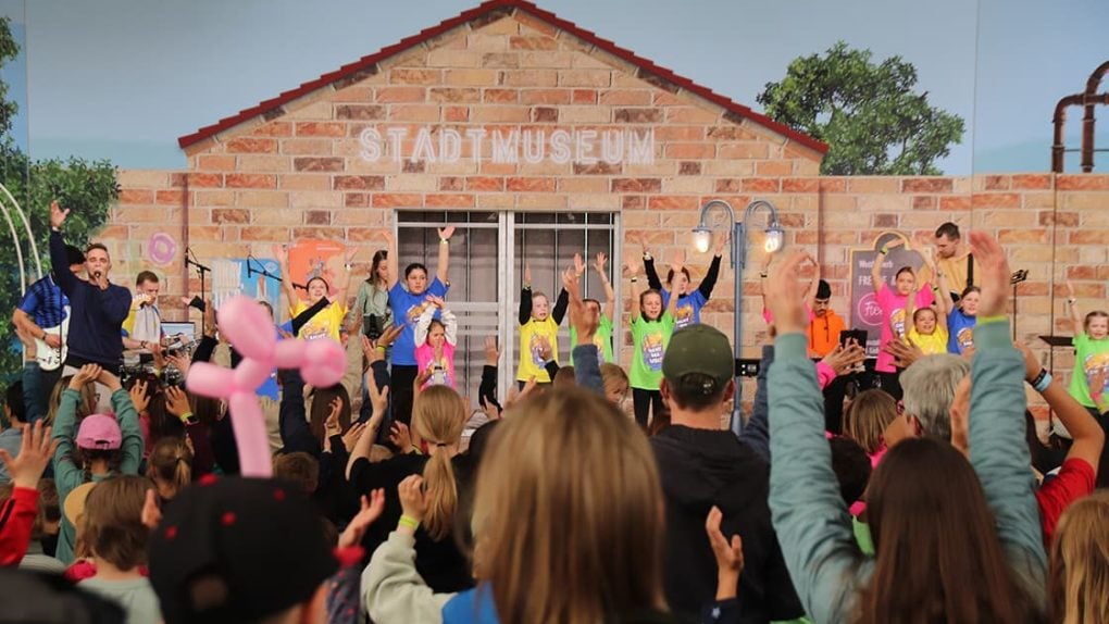 Eine Gruppe von Kindern spielt energisch auf der Bühne, während eine Menschenmenge vor einem Gebäude mit der Aufschrift "Stadtmuseum" zusieht.