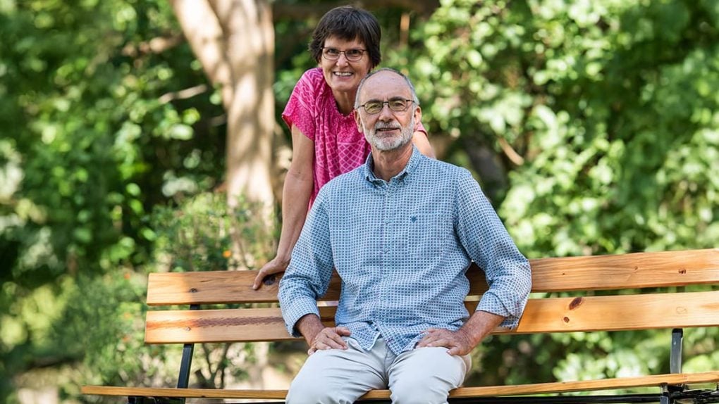 Eine Frau steht hinter einem sitzenden Mann auf einer Holzbank im Freien, beide lächeln inmitten von Grünzeug.