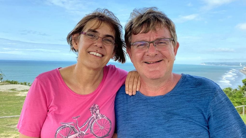Lächelnde Frau und Mann mit Brille posieren zusammen im Freien mit Meer und blauem Himmel im Hintergrund.