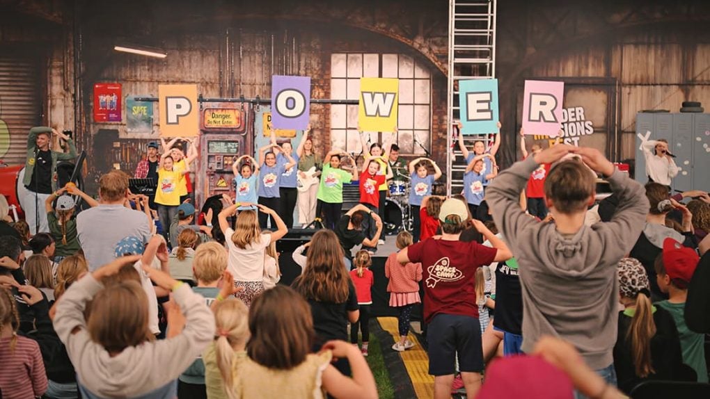 Die Kinder auf der Bühne halten bunte Buchstaben hoch, die POWER buchstabieren, während eine Menge von Kindern ihre Handbewegungen beobachtet und nachmacht.
