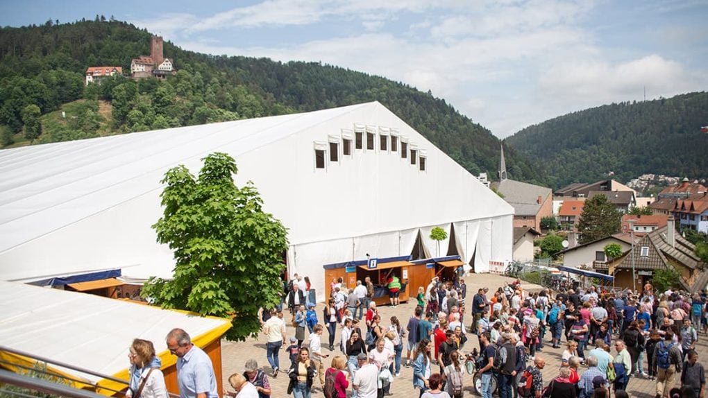 Eine große Menschenmenge versammelt sich vor einem weißen Veranstaltungszelt in einer malerischen, hügeligen Gegend mit Bäumen und entfernten Gebäuden.