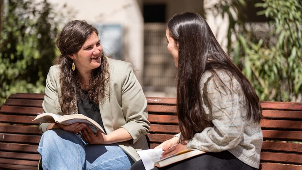 Zwei Frauen sitzen draußen auf einer Bank, lächeln und unterhalten sich, während sie aufgeschlagene Bücher im Schoß halten.