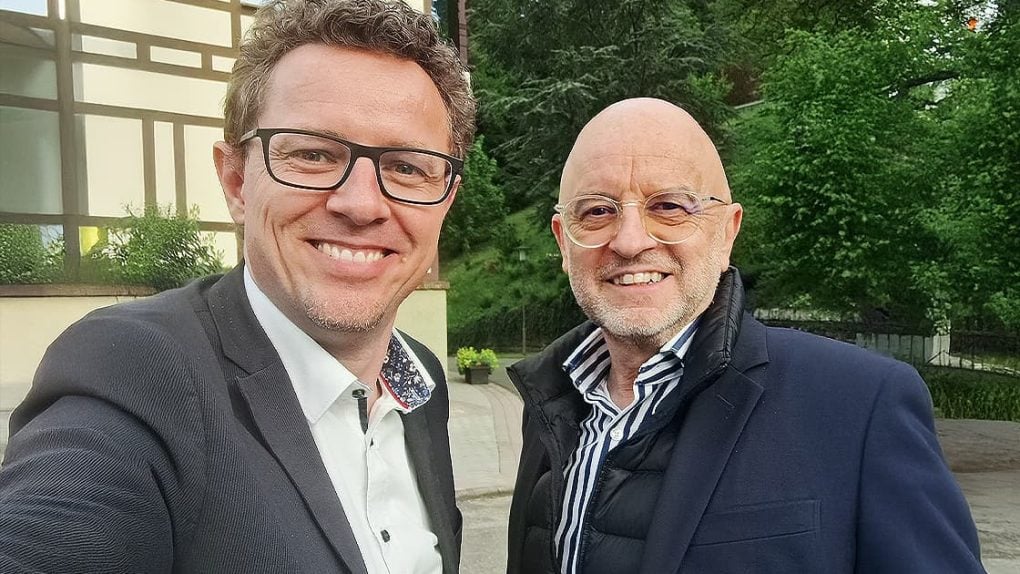 Zwei Männer in Anzügen lächeln im Freien vor einem Gebäude und grünen Bäumen.