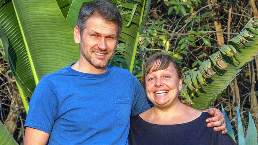Ein Mann und eine Frau lächeln im Freien mit großen grünen Blättern und Bäumen im Hintergrund.