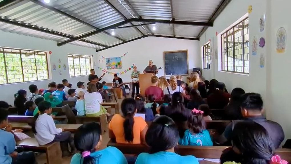 Eine Gruppe von Personen sitzt auf Bänken in einem Klassenzimmer und hört einer Person zu, die an einem Podium neben einer Kreidetafel spricht.