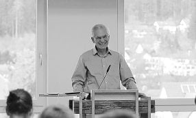 Älterer Mann, der lächelnd auf einem Podium vor einem Publikum spricht, mit großen Fenstern im Hintergrund.