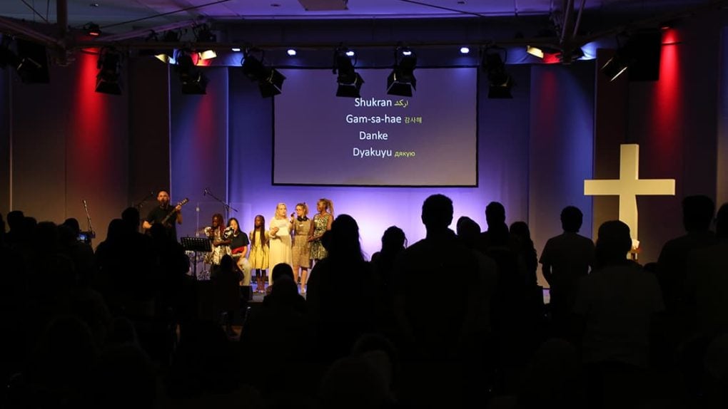 Ein Gottesdienst mit Menschen, die auf der Bühne singen; auf einem Bildschirm über ihnen wird "Danke" in mehreren Sprachen angezeigt.