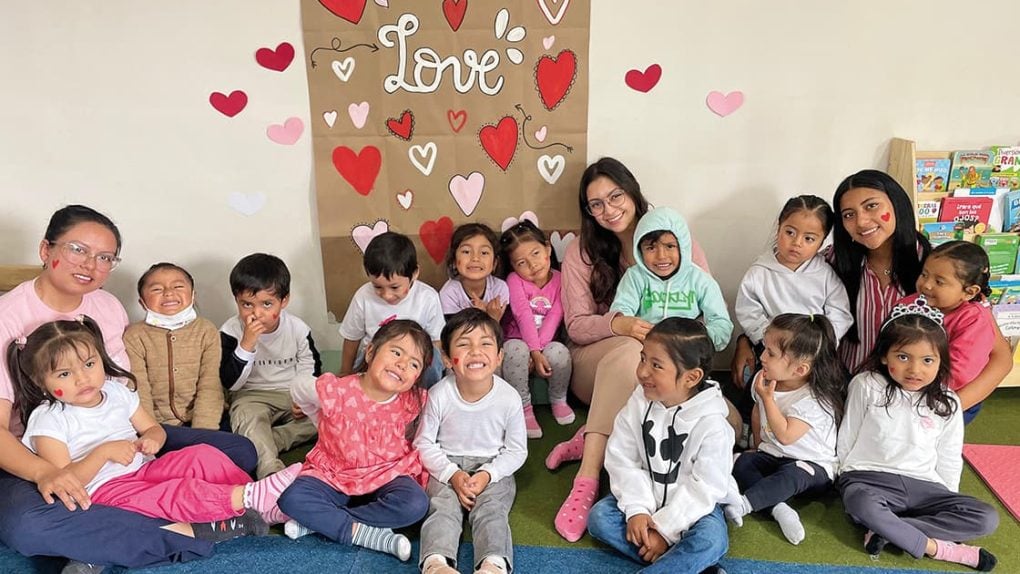 Eine Gruppe kleiner Kinder und Erwachsener sitzt lächelnd vor einer Wand, die mit Herzen und dem Wort „Love“ verziert ist.