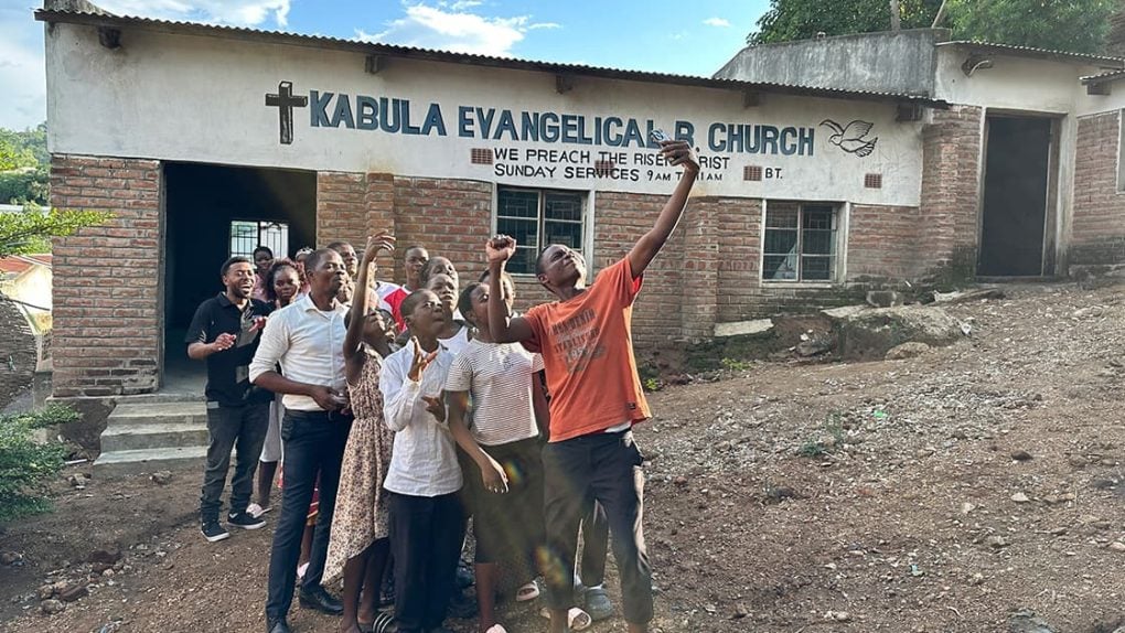 Vor der Kabula Evangelical Church steht eine Gruppe von Menschen, von denen einige die Arme heben und gemeinsam nach oben blicken.