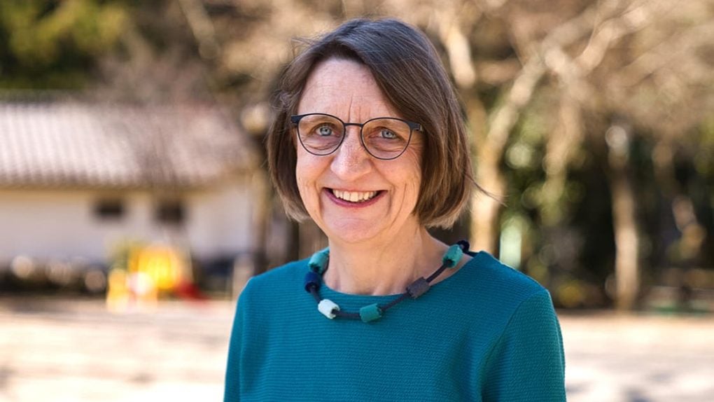 Eine lächelnde Frau mit kurzen braunen Haaren und Brille, die ein blaugrünes Oberteil trägt, steht im Freien vor einem Hintergrund aus Bäumen.