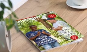 Die Zeitschrift „Mission Weltweit“ liegt auf einem Tisch; darauf sind zwei Männer zu sehen, die sich im Freien unterhalten, einer davon in traditioneller Tracht.