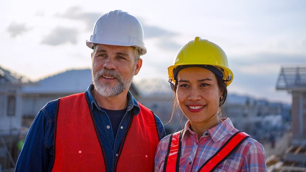 Zwei Bauarbeiter, die Sicherheitswesten und Schutzhelme tragen, stehen draußen auf einer Baustelle und lächeln in die Kamera.