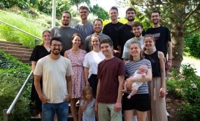 Eine Gruppe aus dreizehn Erwachsenen und zwei Kindern, die im Freien inmitten grüner Bäume und Pflanzen posieren und lächeln.