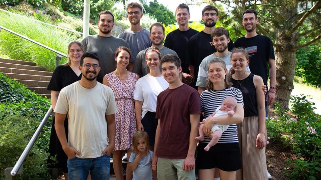 Eine Gruppe aus dreizehn Erwachsenen und zwei Kindern, die im Freien inmitten grüner Bäume und Pflanzen posieren und lächeln.