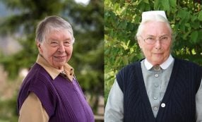 Zwei ältere Frauen, die beide im Freien lächeln; die eine trägt eine lila Weste, die andere eine marineblaue Weste und eine weiße Mütze.