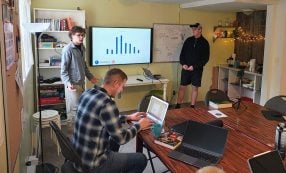 Zwei Personen zeigen ein Balkendiagramm auf einem Bildschirm, während eine weitere Person an einem Tisch in einem Büro auf einem Laptop tippt.