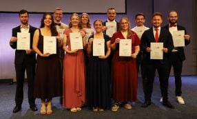 Elf Personen in festlicher Kleidung stehen gemeinsam auf einer Bühne bei einer Veranstaltung, lächeln und halten Urkunden in den Händen.