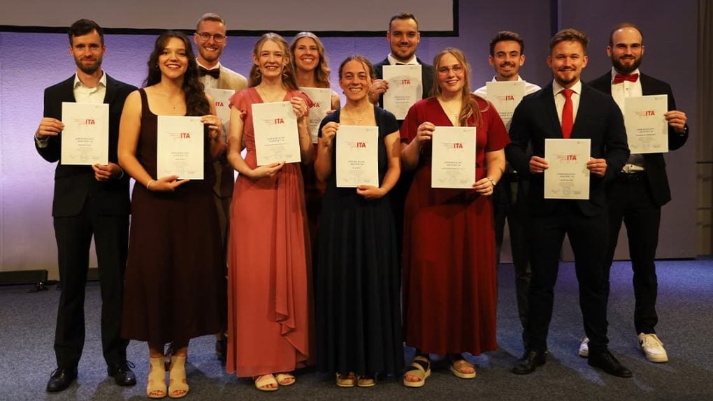 Elf Personen in festlicher Kleidung stehen gemeinsam auf einer Bühne bei einer Veranstaltung, lächeln und halten Urkunden in den Händen.