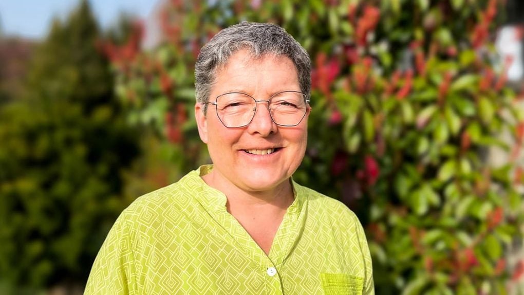 Eine Person mit Brille und einem hellgrünen Hemd, die im Freien lächelt, im Hintergrund sind Grünpflanzen und rote Blumen zu sehen.