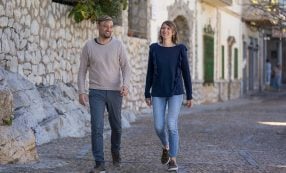 Ein Mann und eine Frau gehen gemeinsam auf einer Kopfsteinpflasterstraße neben einer Steinmauer entlang, lächeln und unterhalten sich.
