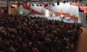 Ein Redner spricht zu einem sitzenden Publikum in einem großen Saal mit einer Bühne, Stühlen und einem „LETS GO!“-Schild.