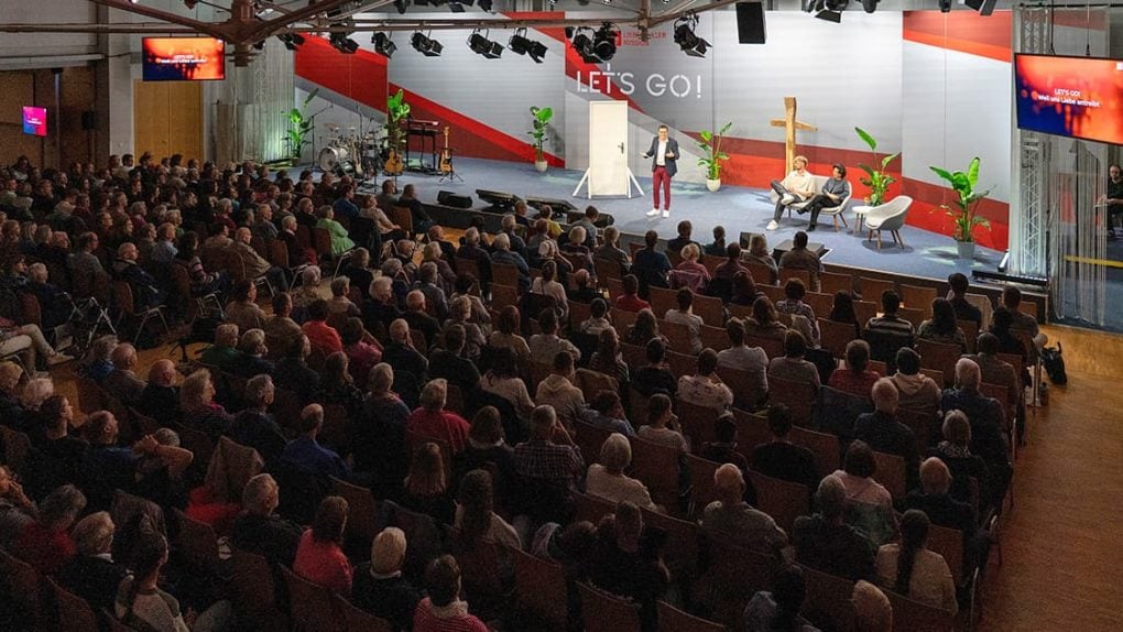 Ein Redner spricht zu einem sitzenden Publikum in einem großen Saal mit einer Bühne, Stühlen und einem „LETS GO!“-Schild.