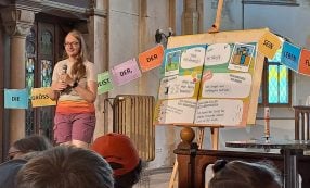 Eine Frau spricht in einer Kirche vor einer bunten Präsentationswand und hängenden Schildern mit deutschen Wörtern.
