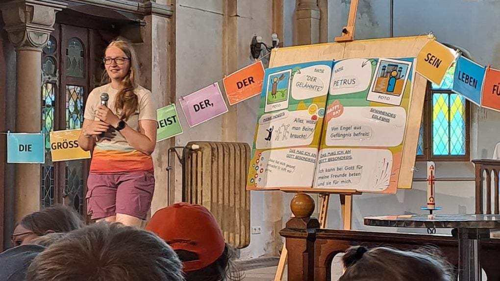 Eine Frau spricht in einer Kirche vor einer bunten Präsentationswand und hängenden Schildern mit deutschen Wörtern.