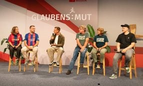 Auf der Bühne sitzen fünf Personen auf Hockern und unterhalten sich, während im Hintergrund ein „Glaubens:Mut“-Banner zu sehen ist.