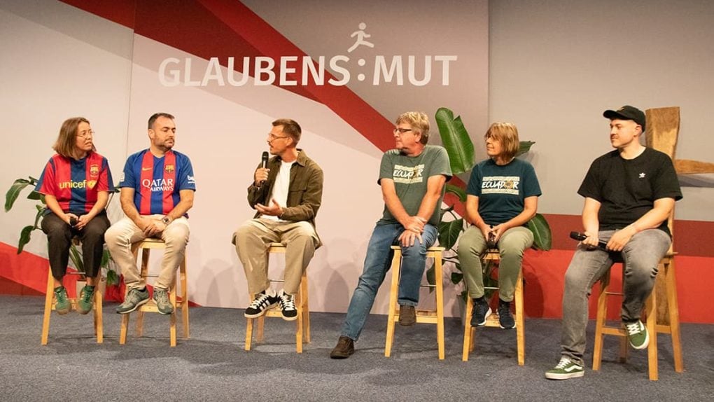 Auf der Bühne sitzen fünf Personen auf Hockern und unterhalten sich, während im Hintergrund ein „Glaubens:Mut“-Banner zu sehen ist.