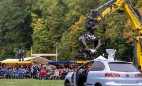 Eine große Menschenmenge sieht zu, wie ein gelber Bagger bei einer Veranstaltung im Freien in der Nähe eines Waldes ein weißes Auto zermalmt.