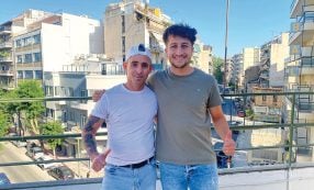 Zwei Männer stehen auf einem Balkon, lächeln in die Kamera, im Hintergrund sind Stadtgebäude zu sehen.