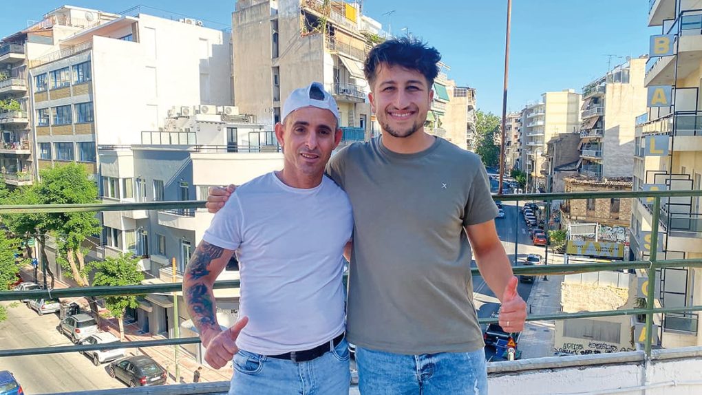 Zwei Männer stehen auf einem Balkon, lächeln in die Kamera, im Hintergrund sind Stadtgebäude zu sehen.