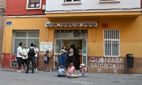 Vor einem gelben Gebäude mit der Aufschrift „Misión Evangélica Urbana“ stehen und sitzen Menschen. Kinder sitzen auf dem Bürgersteig.